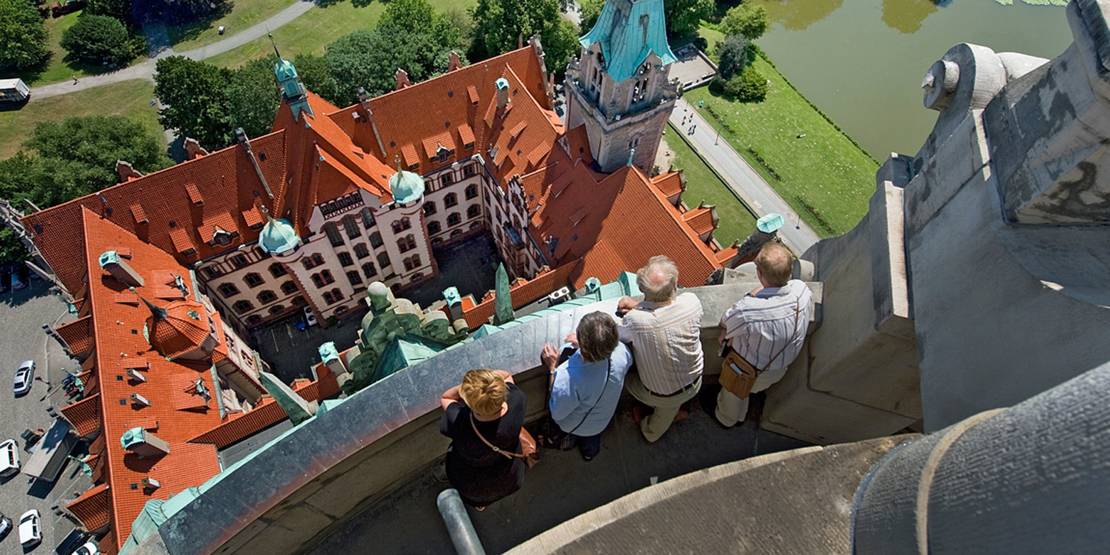 Blick von oben auf vier Personen, die auf der Aussichtsplattform des Rathausturms nach unten auf das Neue Rathaus blicken.